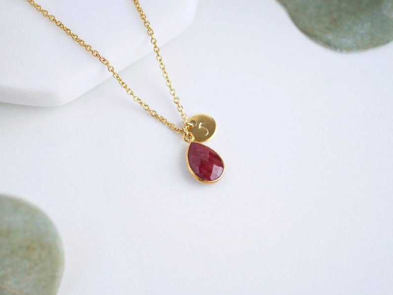 May include: A gold chain necklace with a teardrop-shaped red gemstone pendant and a small gold disc charm with the number "5" engraved on it.