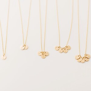 May include: Gold-colored necklaces with delicate chains and small, round pendants. Some pendants feature single initials, while others display multiple initials. The necklaces are displayed against a white background, highlighting the jewelry's simple elegance.