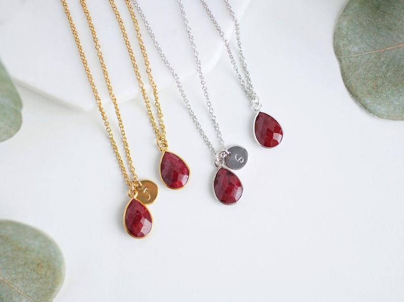May include: Three gold and silver necklaces with red gemstone teardrop pendants and initial charms.