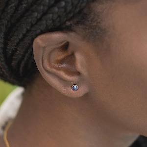 May include: A small, round stud earring featuring a deep blue gemstone set in a simple metal bezel. The earring is positioned centrally on the earlobe, showcasing a minimalist design with a polished finish and a secure, circular metal setting.