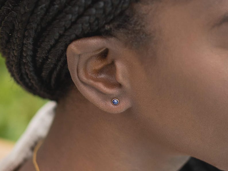 May include: A small, round stud earring featuring a deep blue gemstone set in a simple metal bezel. The earring is positioned centrally on the earlobe, showcasing a minimalist design with a polished finish and a secure, circular metal setting.