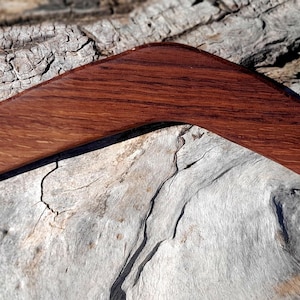 Op de afbeelding: Een houten boemerang met een gebogen, hoekig ontwerp. De boemerang is gemaakt van donkerbruin hout met zichtbare nerfpatronen. Het ligt op een gestructureerd, grijs en bruin oppervlak, waarschijnlijk hout of schors.