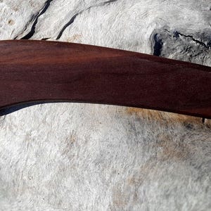 Può includere: Un attrezzo da massaggio in legno curvo, di colore marrone scuro. Lo strumento ha un design liscio ed ergonomico, ideale per applicare pressione sui muscoli. La venatura del legno è visibile, aggiungendo alla sua estetica naturale. Progettato per l'automassaggio e il relax, circa 25 cm di lunghezza.