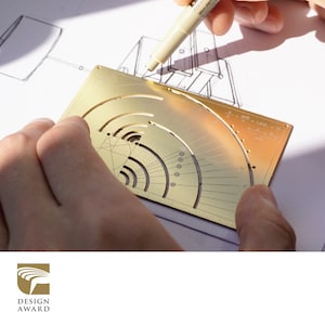 May include: A gold metal protractor with a circular scale and a ruler on the bottom edge. The protractor is being used to draw a line on a piece of paper.