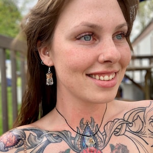 Könnte beinhalten: Eine Frau mit einem Nasenring und Tattoos trägt eine Halskette und Ohrringe mit kleinen Glasflaschen, die mit getrockneten Blumen und Kristallen gefüllt sind.