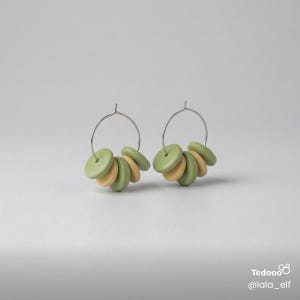 May include: A pair of hoop earrings with silver-colored hoops. Each earring features a cluster of green and tan-colored circular beads. The earrings are displayed against a plain, light gray background. The earrings are a type of jewelry.