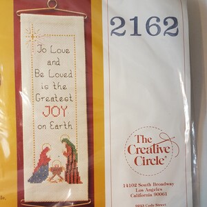 May include: Cross-stitch kit with a religious theme, displaying the text "To Love and Be Loved is the Greatest JOY on Earth." The kit includes a needlepoint banner with a gold-coloured hanging rod and a small bell.