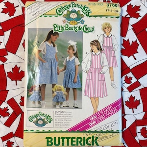 May include: A vintage sewing pattern for Cabbage Patch Kids dolls. The pattern is for a dress with an apron overlay. The pattern is from the 1980s and features a pink and white striped dress and a blue and white floral dress. The pattern is for sizes 7-8-10.