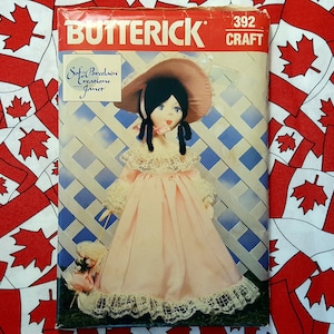 May include: A vintage sewing pattern for a porcelain doll, featuring a pink dress with lace trim and a matching hat. The pattern is from Butterick and is labelled Craft 392. The doll is holding a small bouquet of flowers.