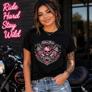 May include: Black t-shirt with a graphic featuring a pink skull wearing a bow, roses, pistons, and the words "Biker Babe." The shirt is worn by a person in blue jeans, with a motorcycle in the background and neon sign that says "Ride Hard Stay Wild."