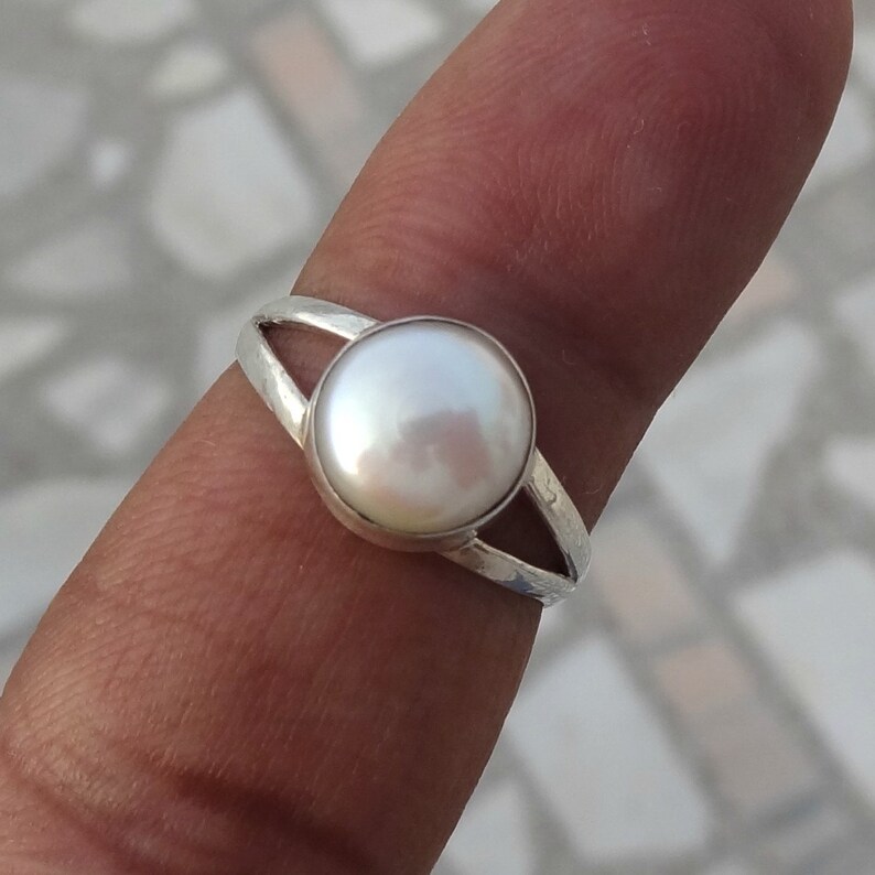 May include: A silver ring featuring a single round, white, iridescent pearl set in a smooth bezel. The band of the ring is split into two thin, textured silver segments that meet at the base, creating a simple and clean design for the jewelry piece.