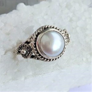 May include: A silver ring featuring a central round white pearl set in a bezel. The band and setting are decorated with a twisted rope texture and small silver bead accents on the sides, creating a detailed metallic frame around the pearl.