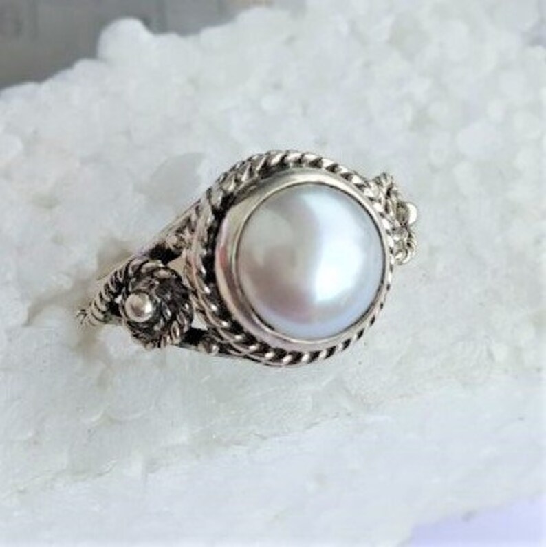 May include: A silver ring featuring a central round white pearl set in a bezel. The band and setting are decorated with a twisted rope texture and small silver bead accents on the sides, creating a detailed metallic frame around the pearl.