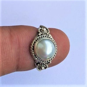 May include: A silver ring featuring a central round white pearl set in a bezel. The band and setting are decorated with a twisted rope texture and small silver bead accents at the top and bottom of the pearl, creating a detailed metal frame.