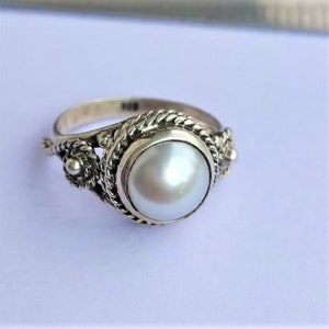 May include: A silver ring featuring a central round white pearl set in a twisted rope-style metal border. The band includes decorative filigree elements on both sides of the setting. The interior of the band is stamped with the number 925.