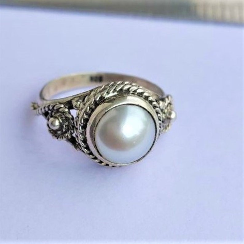 May include: A silver ring featuring a central round white pearl set in a twisted rope-style metal border. The band includes decorative filigree elements on both sides of the setting. The interior of the band is stamped with the number 925.