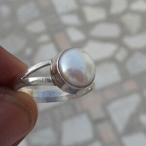 May include: A silver ring featuring a single, round, white pearl set in a smooth bezel. The band of the ring is split into two thin, polished silver segments that meet at the base, creating a simple and clean design for this piece of jewelry.