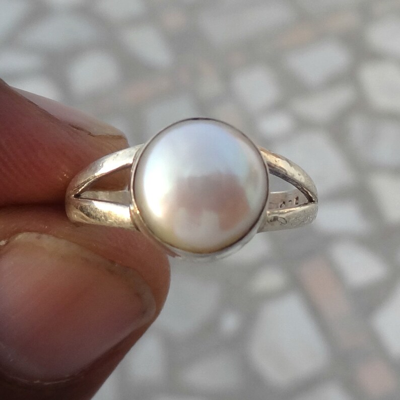 May include: A silver ring featuring a single, round, white pearl set in the center. The band of the ring has a split design on both sides, leading up to the circular setting that holds the pearl securely in place with a smooth, polished finish.