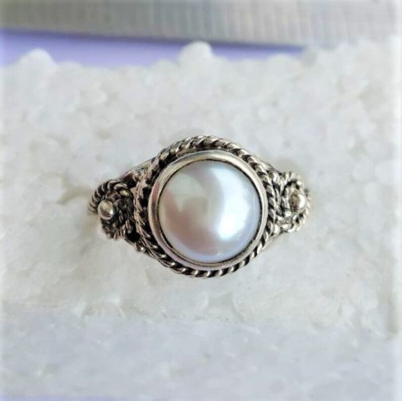 May include: A silver ring featuring a central round white pearl set in a bezel. The band is decorated with a twisted rope pattern and small metal spheres on each side of the setting, creating a detailed texture around the smooth pearl surface.