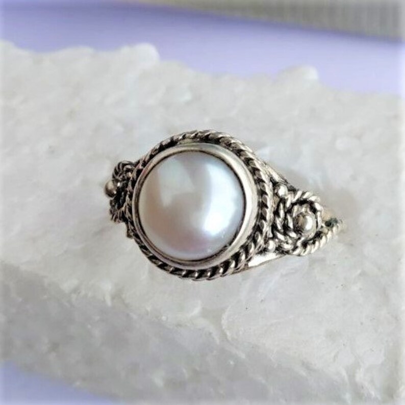 May include: A silver ring featuring a round, iridescent white pearl set in a bezel. The band and setting are decorated with a twisted rope texture and small metal spheres, creating a detailed, textured appearance around the central pearl.