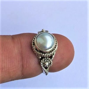 May include: A silver ring featuring a central round white pearl set in a smooth bezel. The band is crafted with a twisted rope texture and includes a decorative knot detail at the base of the setting, creating a detailed metallic finish.