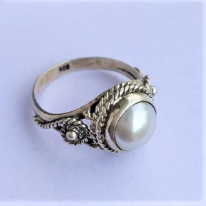 May include: A sterling silver ring featuring a central round white pearl set in a bezel. The band is decorated with a twisted rope pattern and small floral accents on the sides. The interior of the band is clearly stamped with the hallmark 925.