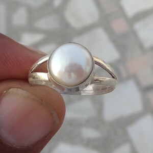 May include: A silver ring featuring a single, round, white pearl set in a smooth bezel. The band of the ring is split into two thin, polished silver segments that meet at the base, creating a simple and clean design for this piece of jewelry.