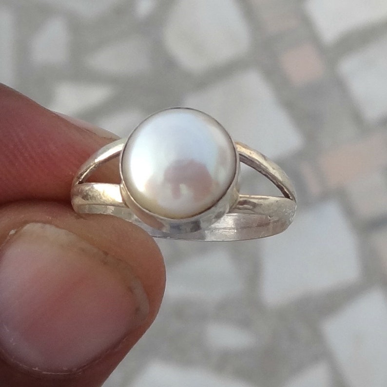 May include: A silver ring featuring a single, round, white pearl set in a smooth bezel. The band of the ring is split into two thin, polished silver segments that meet at the base, creating a simple and clean design for this piece of jewelry.
