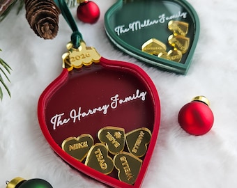 May include: Two teardrop-shaped Christmas ornaments, one red and one dark green, featuring the text The Harvey Family and The Meller Family. Each ornament contains small gold heart-shaped charms with names engraved, and the year 2026 is etched on the gold-coloured top cap.