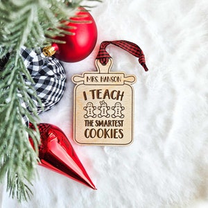 May include: Wooden Christmas bauble with a red and black ribbon. The bauble is shaped like a cookie jar and says "Mrs. Hanson I Teach The Smartest Cookies".