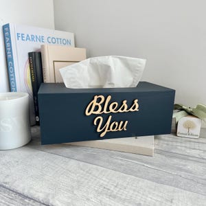 May include: A dark blue tissue box with the words "Bless You" in wooden lettering. The box contains white tissues and is placed on a light grey surface. Books and a candle are visible in the background.