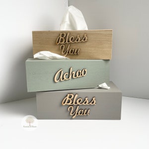 May include: Three wooden tissue box covers stacked on top of each other. The top cover is natural wood with the words "Bless You" written in cursive on it. The middle cover is painted a light green with the word "Achoo" written in cursive on it. The bottom cover is painted a light grey with the words "Bless You" written in cursive on it.