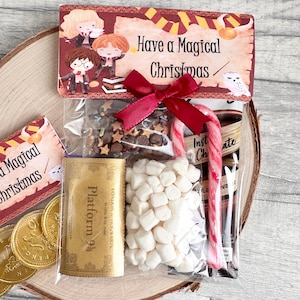 May include: A festive cellophane gift bag containing hot chocolate ingredients, marshmallows, and a candy cane. The bag is adorned with a red ribbon and a tag featuring the text "Have a Magical Christmas" and cartoon characters, perfect for the holidays.