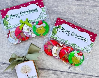 Może przedstawiać: Dwie przezroczyste plastikowe torebki wypełnione czekoladowymi monetami z motywem Grincha. Torebki mają czerwono-zielone etykiety z napisem "Merry Grinchmas". Na pierwszym planie znajduje się małe drewniane pudełko z wzorem drzewa i tekstem "Rosewood & Blossom".