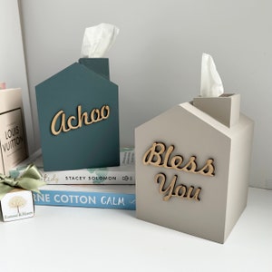 May include: Two house-shaped tissue box holders. One is teal with the word "Achoo" in wooden letters, and the other is light grey with "Bless You." Both have tissue emerging from the chimney.