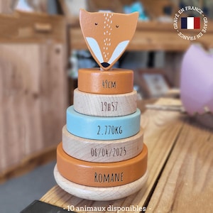 Peut inclure: Un jouet d'empilage en bois avec une tête de renard. Il comporte des anneaux orange, bleus et en bois naturel avec des détails gravés, dont des mesures et le nom "Romane". Le texte "10 animaux disponibles" est en bas. Fabriqué en France.