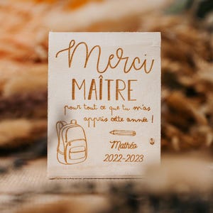 Peut inclure: Plaque en bois rectangulaire gravée avec les mots "Merci MAÎTRE" en écriture cursive. Le texte "pour tout ce que tu m'as appris cette année!" est écrit en dessous. Un sac à dos et un crayon sont également gravés. Le nom "Mathéo" et l'année "2022-2023" sont en bas.