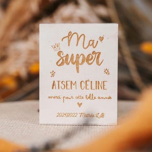 Peut inclure: Un bloc de bois clair avec texte gravé et éléments décoratifs. Le texte dit "Ma super ATSEM Céline merci pour cette belle année 2021/2022 Mathéo L.B." avec une couronne, des cœurs et des étoiles.