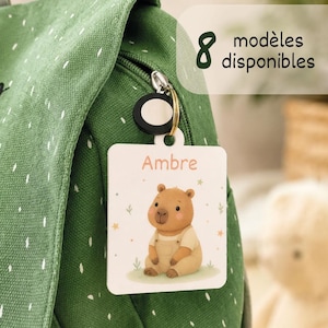 Peut inclure: Une étiquette de sac rectangulaire blanche attachée à un sac à dos en toile verte avec un anneau métallique. L'étiquette présente un capybara en dessin animé portant une salopette beige, le prénom Ambre et 8 modèles disponibles.