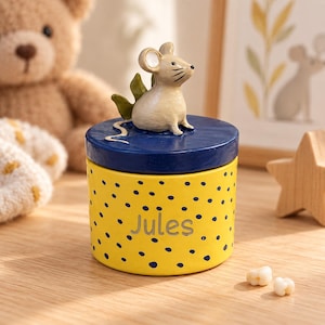 Peut inclure: Un récipient cylindrique jaune à pois bleus avec le prénom Jules imprimé en gris. Le couvercle est bleu foncé et présente une figurine de souris beige sculptée avec des feuilles vertes derrière, posée sur la surface plane du couvercle.