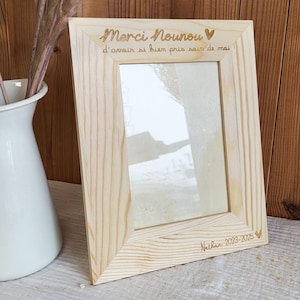 Peut inclure: Un cadre photo en bois clair avec texte gravé. Le cadre est de forme rectangulaire avec une vitre transparente. Le texte comprend "Merci Nounou" et "Nathan 2023-2025". Le cadre est posé contre un vase blanc et un fond en bois.