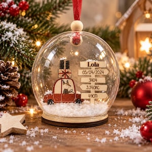 Peut inclure: Une boule de Noël en verre transparent contenant une voiture rouge avec un cadeau sur le toit, un panneau en bois avec le texte Lola, 05/04/2024, 3H04, 3.340KG et 49CM, posé sur une base enneigée avec un ruban rouge et une perle en bois.