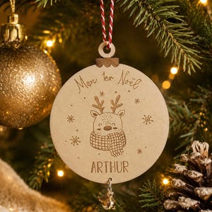 Peut inclure: Une décoration de Noël ronde en bois gravée d'un renne portant une écharpe, entouré de flocons de neige. Le texte indique Mon 1er Noël et ARTHUR. Elle est suspendue par une ficelle rayée rouge et blanche et comporte une petite clochette en métal.