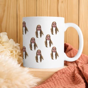 Peut inclure: Mug en céramique blanche avec un motif répété d'un visage. Les visages sont espacés uniformément sur la surface de la tasse. La tasse a une grande anse incurvée et repose sur une surface en bois.