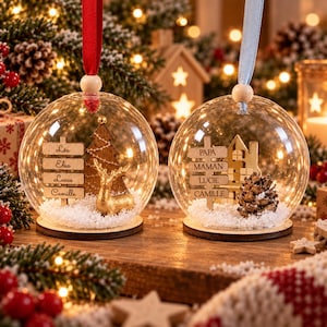 Peut inclure: Deux boules de No&euml;l en verre transparent avec socles en bois et rubans. La boule de gauche contient un cerf dor&eacute;, un sapin en bois et un panneau avec les noms L&eacute;a, Elsa, Lucas, Camille. La boule de droite contient une pomme de pin, une maison en bois et un panneau avec PAPA, MAMAN, LUCIE, CAMILLE.