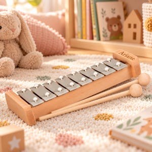 Peut inclure: Un xylophone en bois doté de huit touches en métal argenté, chacune munie d'un petit bouton blanc. Le cadre en bois porte le nom Ariane gravé. Deux maillets en bois à tête ronde sont placés le long de l'instrument sur la base.