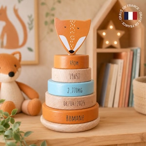 Peut inclure: Un jouet d'empilage en bois avec une tête de renard et des blocs circulaires. Les blocs sont orange, bleu et en bois naturel, avec des mesures et un nom imprimés. Le jouet est conçu pour les enfants.