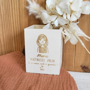 Peut inclure: Un coffret en bois blanc avec un dessin de femme et le texte "Merci Maîtresse Julia de m'avoir aidé à grandir !" et le nom "Milo". Le coffret est décoré d'un dessin de femme.