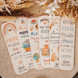Peut inclure: Une collection de marque-pages en bois avec divers motifs et textes. Chaque marque-page présente des illustrations uniques, notamment un arc-en-ciel, des livres et un globe. Le texte comprend des noms, des dates et le mot "Merci".