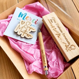 Op de afbeelding: Een geschenkdoos met een houten pen, een kaart en een houten uitsparing. De pen is lichtbruin met zilveren accenten en de tekst "Super Maîtresse !" De kaart zegt "Merci de mi sandir !" en heeft een houten uitsparing met "Super Maîtresse".